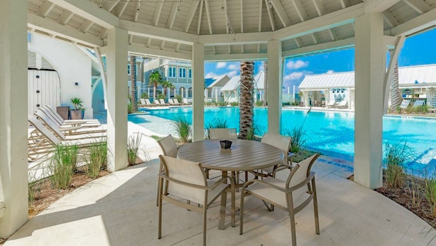 Un espace extérieur aménagé, abrité et doté de sièges, offrant une vue sur la piscine, avec une table ronde et des chaises, ainsi qu’une vue sur la piscine et les bâtiments du complexe hôtelier.