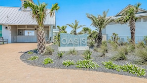 A sign with "OASIS" in front of a building with a metal roof, surrounded by palm trees and landscaped areas. - Shorely the Best | Palmilla Condo with Pool, Dining & Boardwalk to Beach (Port Aransas)
