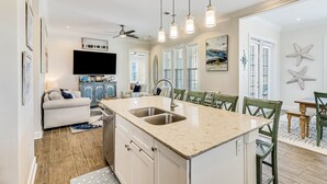 A modern kitchen with a central island, a dining area with green chairs, and a flat-screen TV mounted on the wall. - Shorely the Best | Palmilla Condo with Pool, Dining & Boardwalk to Beach (Port Aransas)