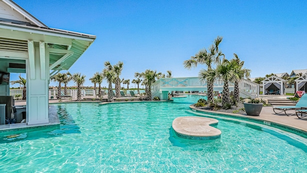 A swimming pool with a heart-shaped island, palm trees, and a covered area with lounge chairs. - Shorely the Best | Palmilla Condo with Pool, Dining & Boardwalk to Beach (Port Aransas)