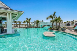 A swimming pool with a heart-shaped island, palm trees, and a covered area with lounge chairs. - Shorely the Best | Palmilla Condo with Pool, Dining & Boardwalk to Beach (Port Aransas)