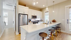 Una cocina moderna con armarios blancos, electrodomésticos de acero inoxidable, una isla grande con taburetes y luces colgantes, con vista al área de estar contigua.