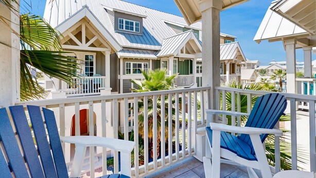 A covered outdoor area with white railings and blue chairs, featuring a view of a building with a gabled roof and multiple windows. - Perfect Palmilla Beach Townhome | Near Resort Pool, Beach Access & Golf (Port Aransas)