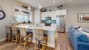 A modern kitchen with a central island, white cabinetry, stainless steel appliances, and a blue sofa in the living area. - Perfect Palmilla Beach Townhome | Near Resort Pool, Beach Access & Golf (Port Aransas)