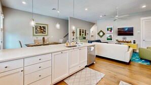A modern kitchen with white cabinets, a built-in oven, and a large island with a sink. There is a white sofa and a green armchair in the living area, and a ceiling fan above. - Upscale townhouse near the pool with bikes, resort boardwalk & golf course (Port Aransas)