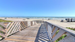Una passerella in legno che conduce a una spiaggia, con veicoli e un cielo azzurro limpido.