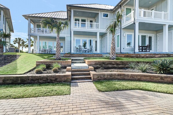 Uma casa de dois andares com varanda frontal, alpendre coberto e jardim ajardinado com palmeiras e arbustos.