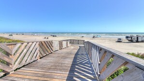 Ein hölzerner Steg führt zu einem Strand mit Fahrzeugen und blauem Himmel.