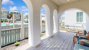 Un espacio al aire libre cubierto con una terraza de madera, columnas blancas y vistas a una piscina y edificios al fondo.