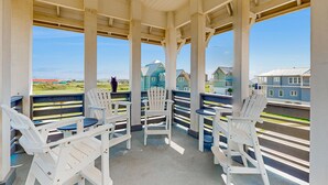 A covered outdoor seating area with white Adirondack chairs and tables, overlooking a residential area with houses and a clear sky.