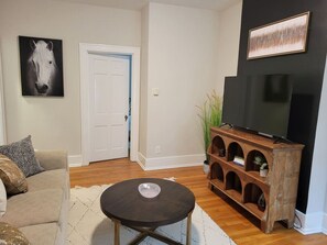 Una sala de estar con un sofá, una mesa de centro, un televisor sobre un mueble de madera y una puerta al fondo.