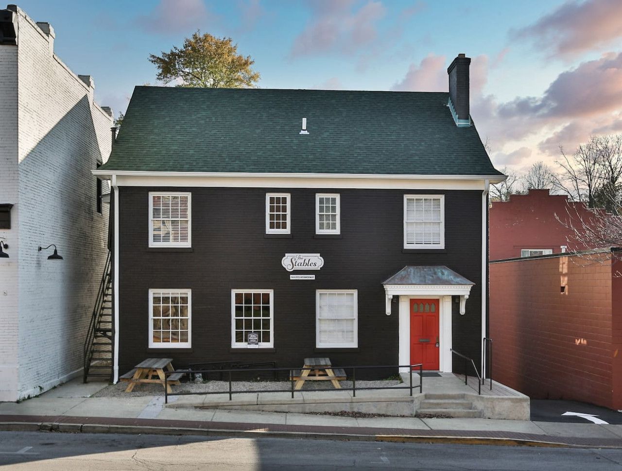 A two-story building with a dark facade, white trim, and a red door. It has multiple windows, a sign reading "The Stables," and a small outdoor seating area with benches.