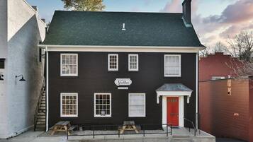 A two-story building with a dark facade, white trim, and a red door. It has multiple windows, a sign reading "The Stables," and a small outdoor seating area with benches.