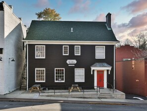 A two-story building with a dark facade, white trim, and a red door. It has multiple windows, a sign reading "The Stables," and a small outdoor seating area with benches. - Downtown Main Street One-Bedroom Suite w/ Hi-Speed Wifi (Shelbyville)