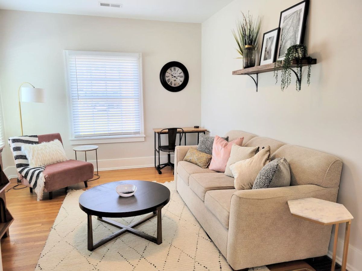Una sala de estar con un sofá color beige, una mesa redonda, un sillón a rayas, una mesita auxiliar, un reloj de pared y una estantería con plantas.