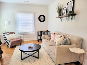 A living room with a beige sofa, a round coffee table, a striped armchair, a small side table, a floor lamp, a clock, and a shelf with plants and pictures. - Downtown Main Street One-Bedroom Suite w/ Hi-Speed Wifi (Shelbyville)