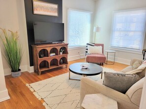 A living room with a flat-screen TV, a wooden entertainment unit, a round coffee table, a beige sofa, and a potted plant. - Downtown Main Street One-Bedroom Suite w/ Hi-Speed Wifi (Shelbyville)