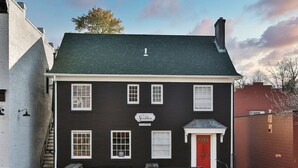 A two-story building with a dark facade, white trim, and a red door. It has multiple windows, a sign reading "The Stables," and a small outdoor seating area with benches. - Downtown Main Street One-Bedroom Suite w/ Hi-Speed Wifi (Shelbyville)