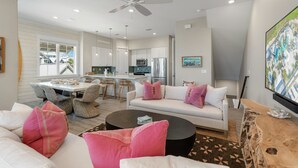 A modern living room with a white sofa, a coffee table, and a dining area with a white table and chairs. - Palmilla Beach | Sunset-View End Unit | Garage & All-Suite | Steps to Pool (Port Aransas)