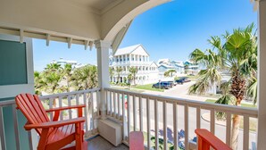 A balcony with two red Adirondack chairs, a white railing, and a view of a residential area with palm trees and houses. - Palmilla Beach | Sunset-View End Unit | Garage & All-Suite | Steps to Pool (Port Aransas)