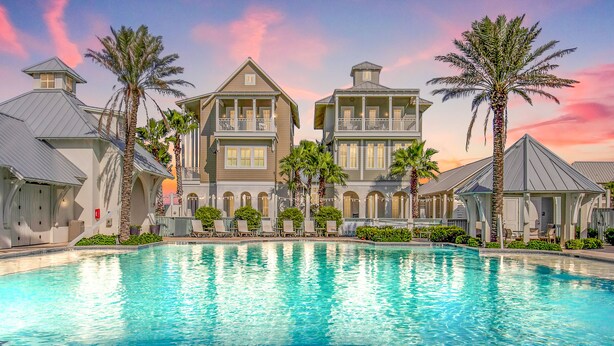 A large swimming pool surrounded by two-story buildings with balconies, palm trees, and a sunset sky. - Luxe Townhome in Palmilla Beach Resort | Beach Service, Pool & Golf Course (Port Aransas)