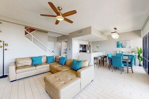 Ein modernes Wohnzimmer mit einer Eckcouch, einem Essbereich mit blauen Stühlen, einem Deckenventilator und einer Küche im Hintergrund.