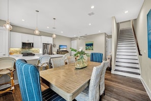 A modern kitchen with white cabinets, a wooden dining table, wicker chairs, and pendant lighting.