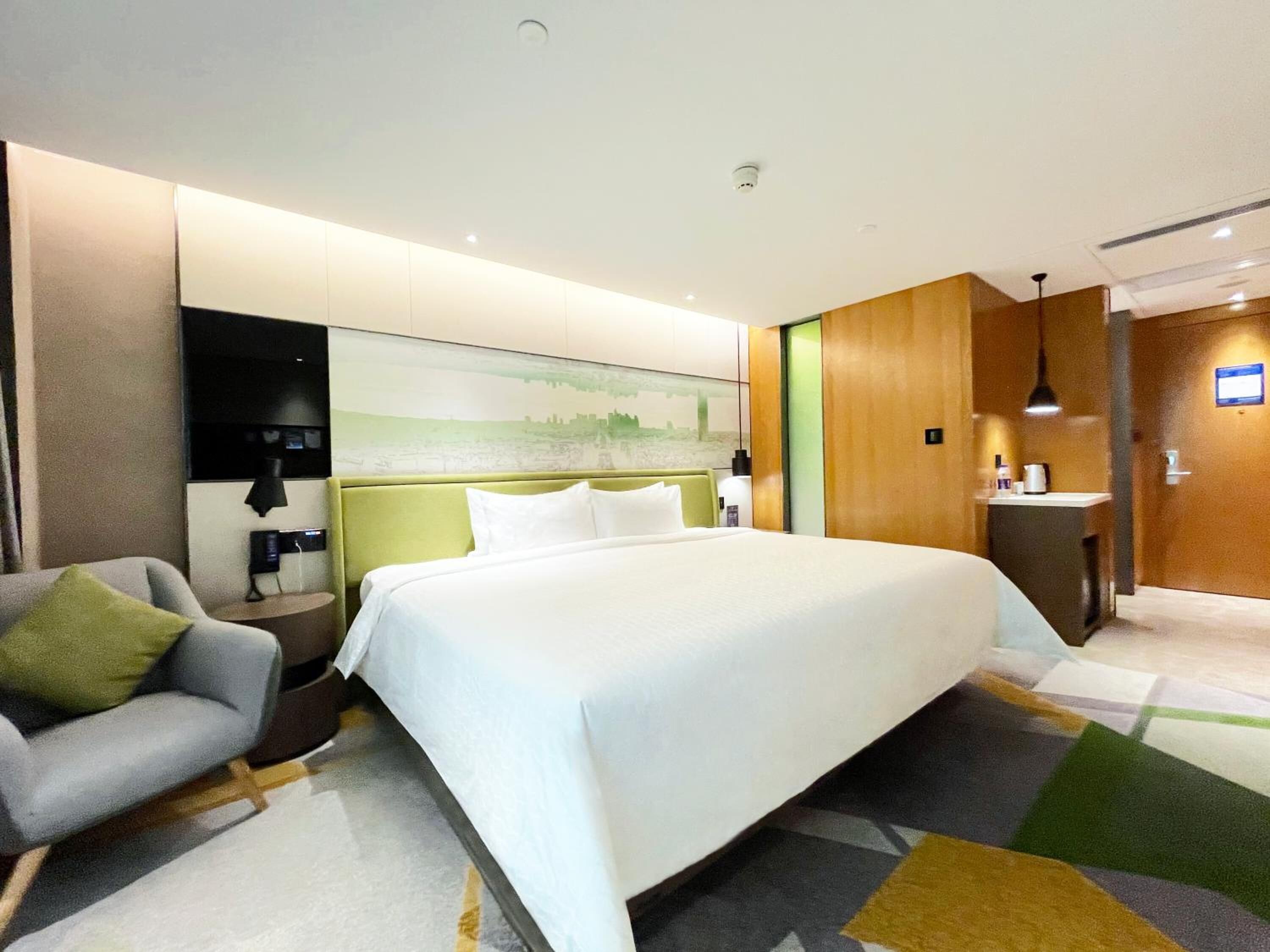A modern hotel room with a large bed, a bedside table with a lamp, a chair, and a view of the cityscape through the window.