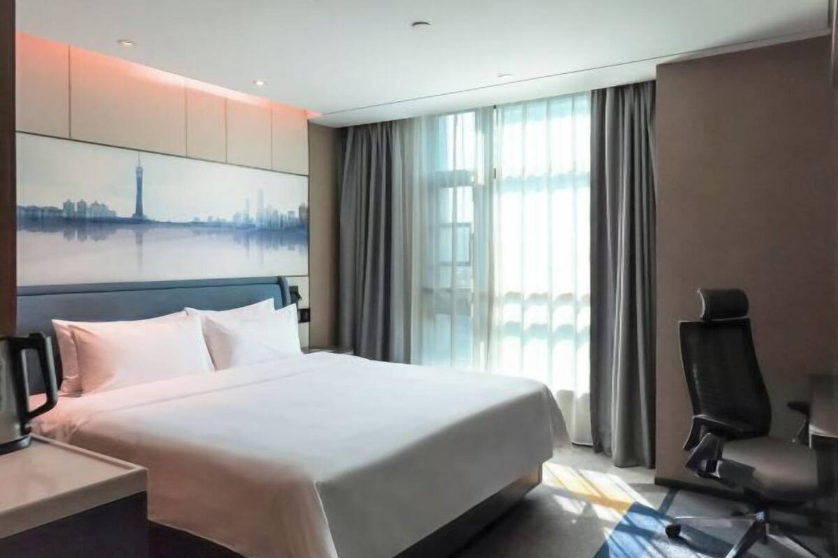 A hotel room with a large bed, a desk with a chair, a painting of a cityscape, and a view of a building through the window.