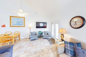 Una sala de estar con televisor, dos sillones estampados, un sofá, una mesa de comedor con sillas, un reloj y un ventilador de techo.