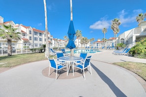 Un espace aménagé au bord de la piscine, avec une table ronde et des chaises bleues, entouré de palmiers et surplombé d’un ciel d’un bleu limpide.