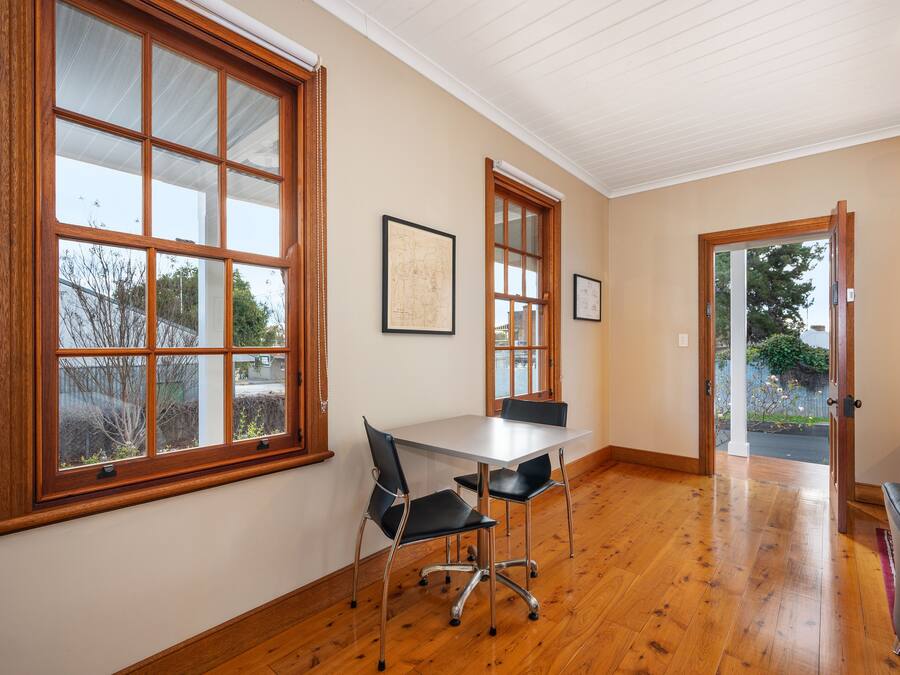 A room with a wooden floor, a white table with two chairs, and large windows with wooden frames.