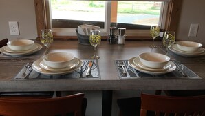 A dining table set with white plates, bowls, and glasses, with a view of a green landscape through a window. - River Rest Luxury Cabin - The Big Hole. Beautiful River and Mountain Views!! (Silver Star)