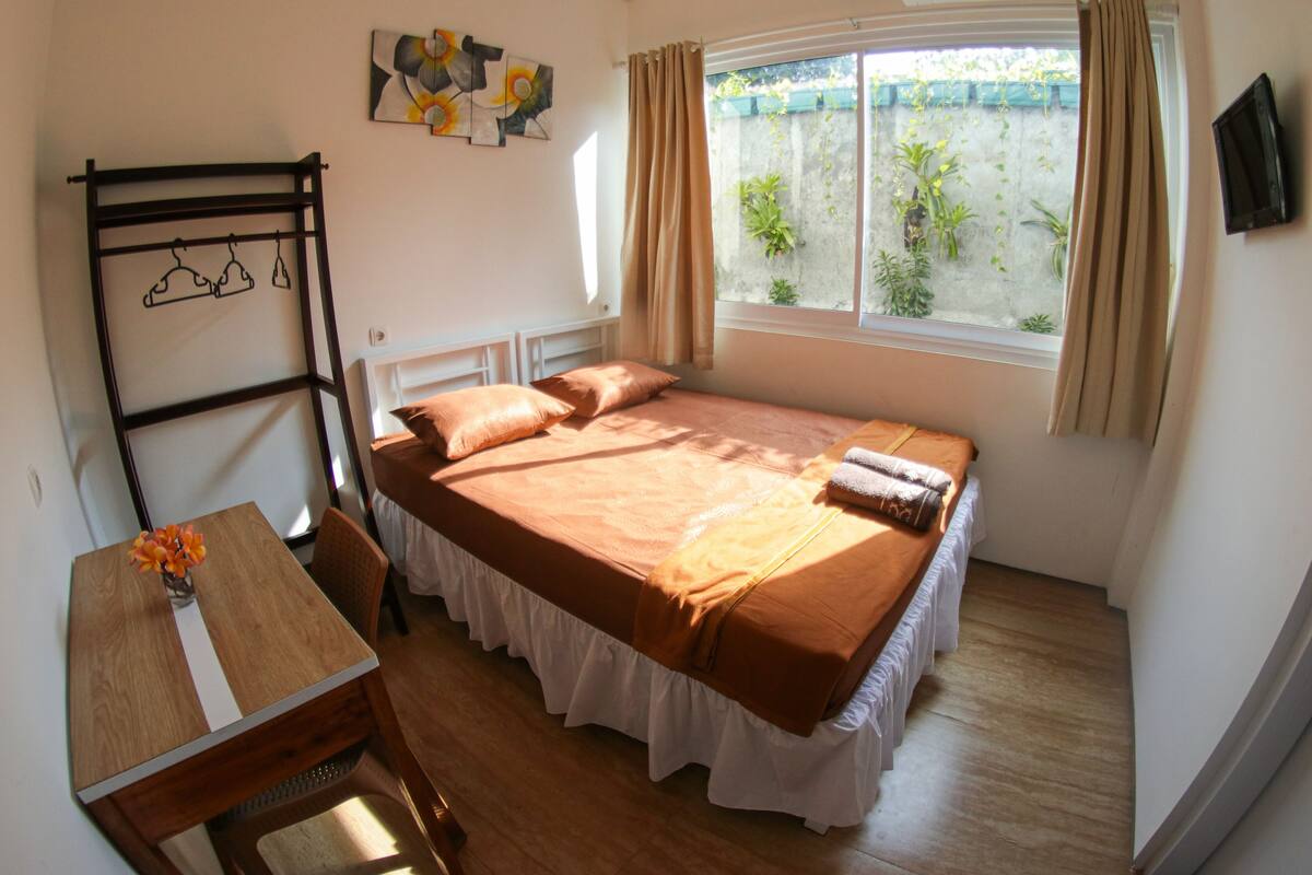 A bedroom with a bed, a desk, a chair, a window with curtains, and a wall-mounted TV.