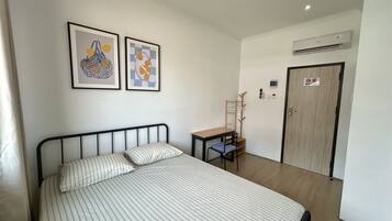 A hotel room with a bed, a desk, a chair, two framed artworks, an air conditioning unit, and a door with a 'No Smoking' sign.
