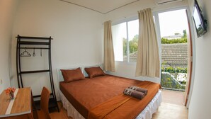 A bedroom with a bed, a desk, and a chair. There is a window with curtains, an air conditioning unit, and a view of greenery outside.