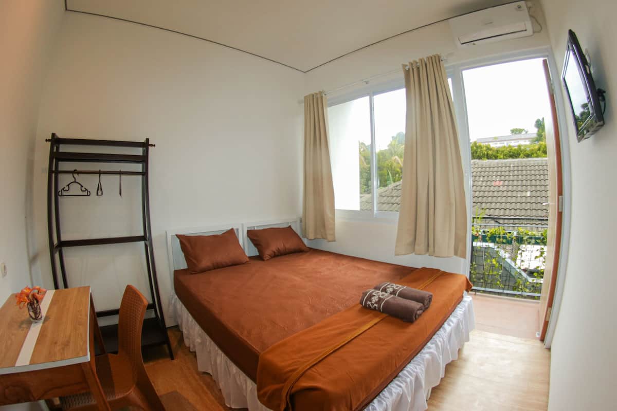 A bedroom with a bed, a desk, and a chair. There is a window with curtains, an air conditioning unit, and a view of greenery outside.