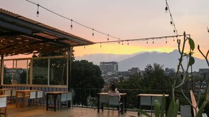 An outdoor seating area with tables and chairs, overlooking a cityscape and mountains during sunset.