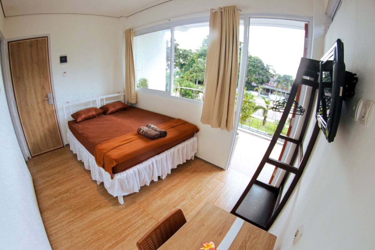 A bedroom with a bed, a small table with a vase of flowers, a TV mounted on the wall, and a window with curtains.