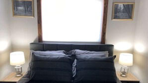 Una cama bien hecha con una colcha oscura a rayas y almohadones, flanqueada por dos mesitas de noche con lámparas, en una habitación con un gran ventanal y cuadros en las paredes.