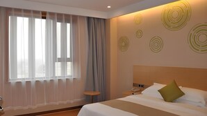 Een hotelkamer met een groot bed, nachtkastje, lamp en raam met dunne gordijnen.