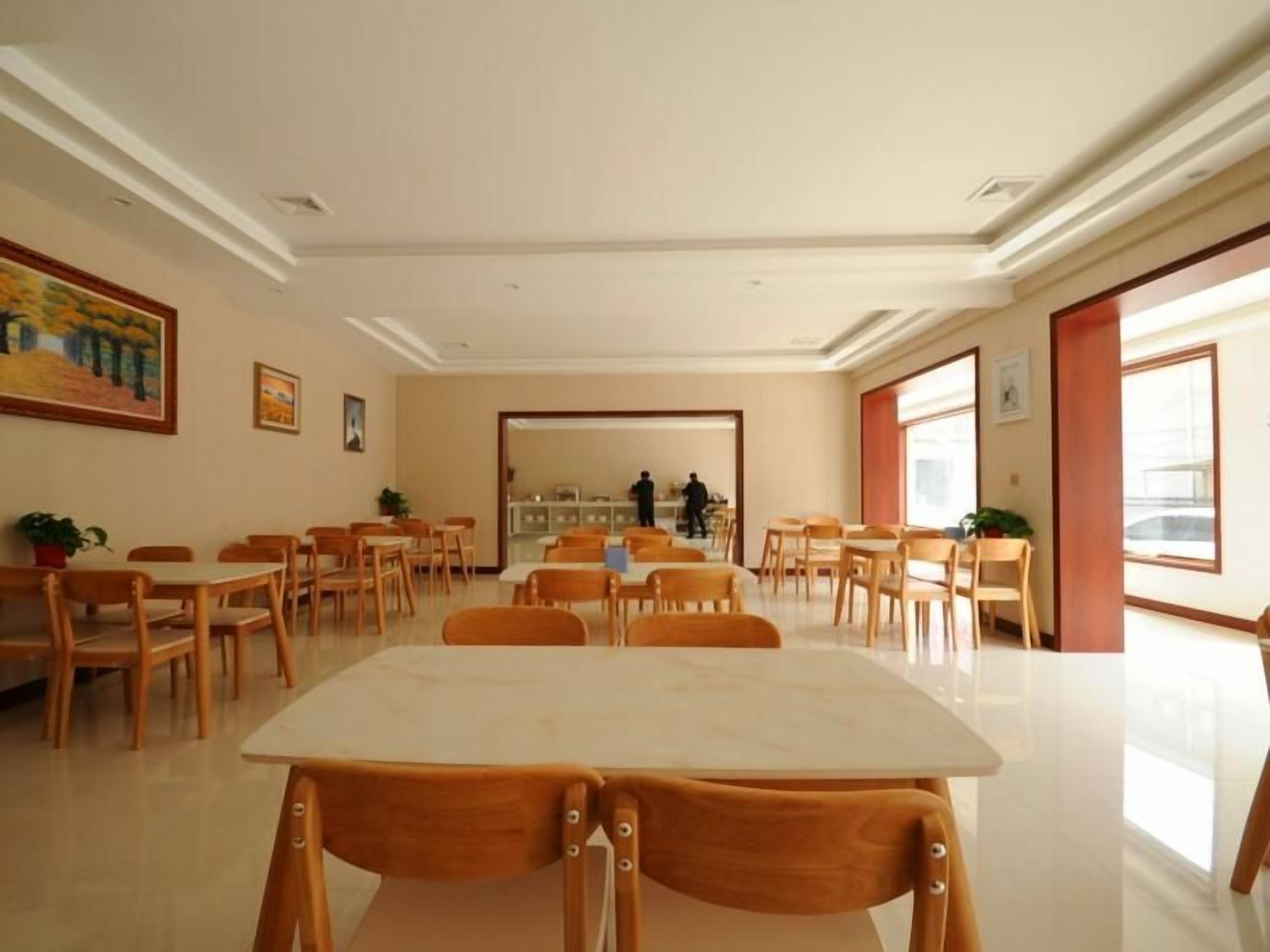 A spacious dining area with wooden tables and chairs, a kitchen in the background, and large windows allowing natural light.