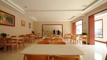 A spacious dining area with wooden tables and chairs, a kitchen in the background, and large windows allowing natural light.