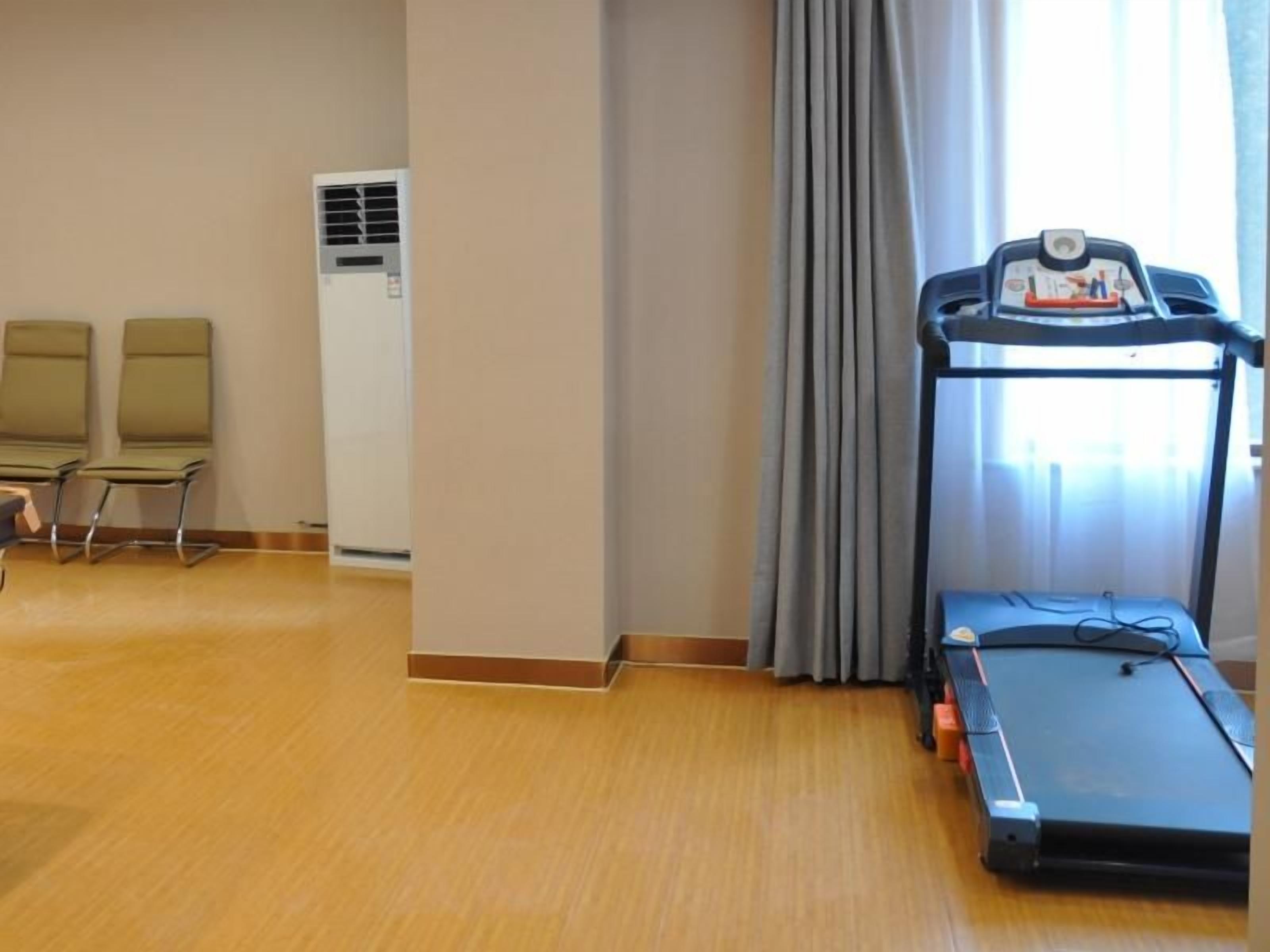 Habitación de hotel con una caminadora, dos sillas, un ventanal con cortinas y un aire acondicionado instalado en la pared.