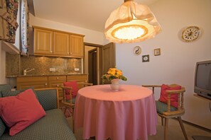 A cozy living room with a round table, a sofa, a television, and a kitchen area in the background.