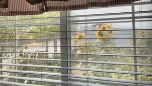 A window with horizontal blinds, a striped curtain, and a view of sunflowers and greenery outside. - The Chick Inn<br>Bed Breakfast and Dinner!!! (Eagle Mountain)