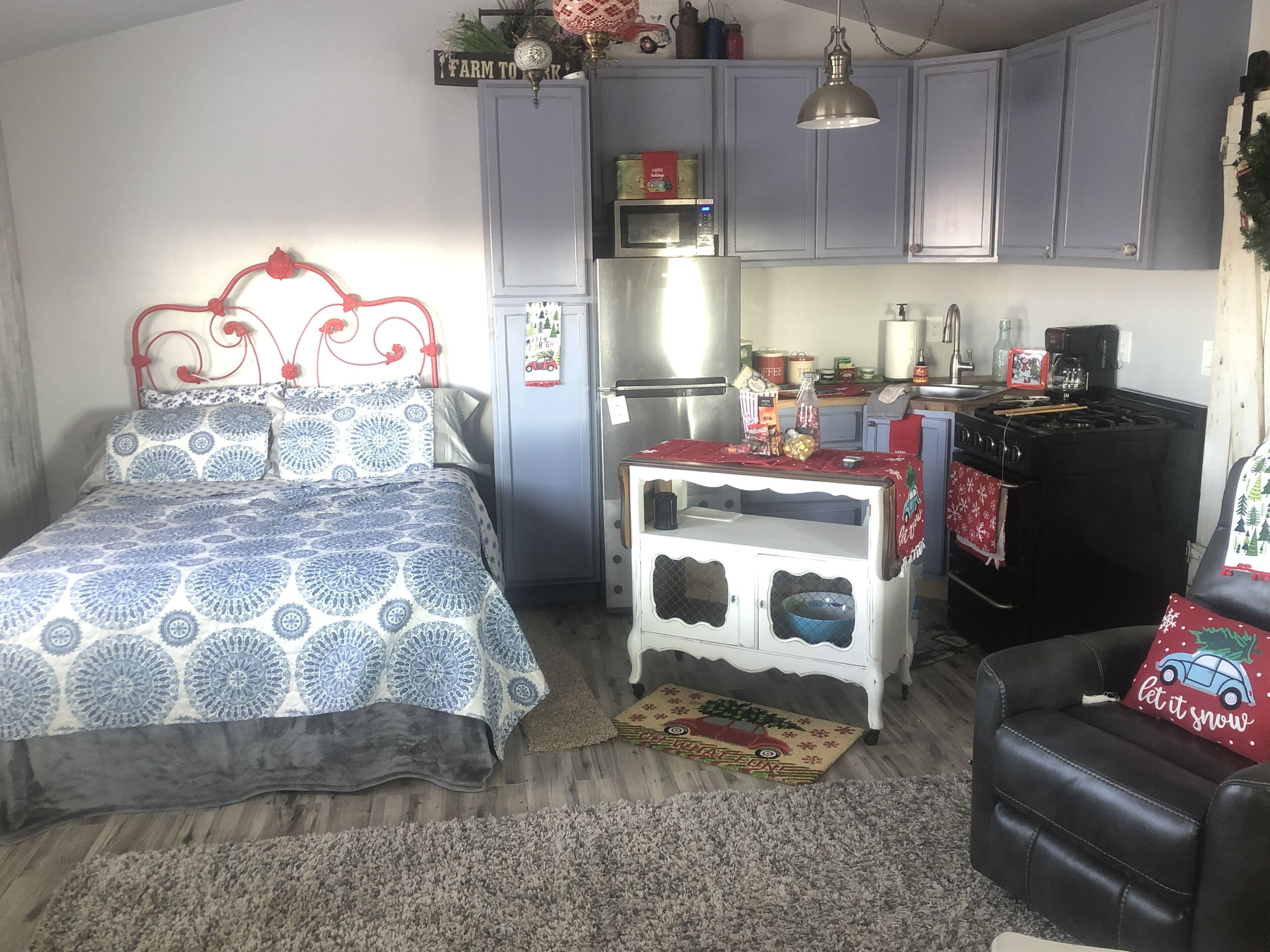 Une chambre à coucher de taille réduite, comprenant un lit, une petite commode blanche, une kitchenette avec une cuisinière et une chaise.