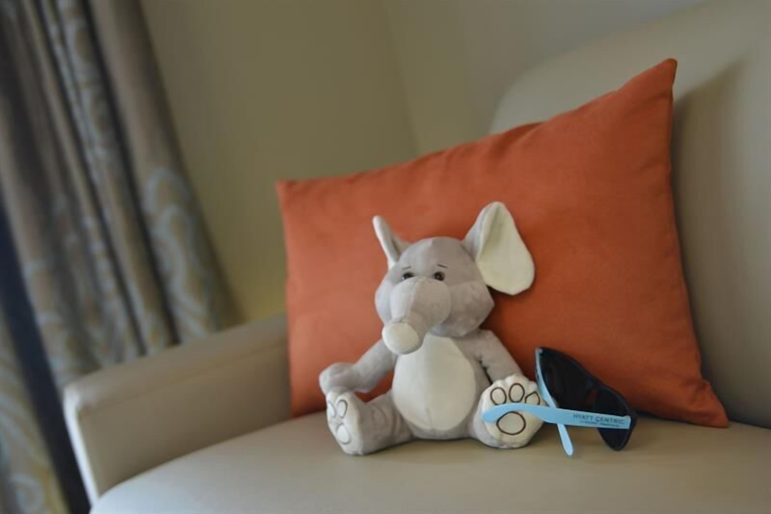A plush toy elephant on a beige sofa with an orange pillow and a pair of sunglasses.
