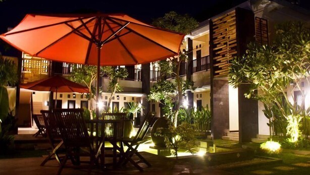 An outdoor seating area with tables and chairs under a large umbrella, surrounded by well-lit buildings and greenery. - Hotel Orizatha (Mataram)