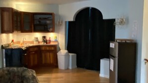 Una sala de estar con un sillón estampado, una cocina con armarios de madera, un refrigerador negro y un cesto de basura.